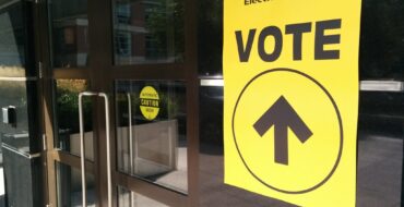 a yellow sign that says "Elections Canada" and "Vote", with an arrow pointing up, on a glass window beside two glass exterior doors on a public building