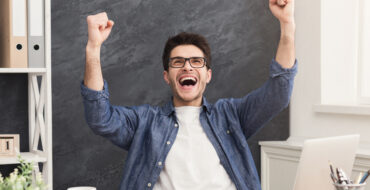man sitting at a desk with a computer, phone, and tablet is leaning back and thrusting his fists triumphantly into the air
