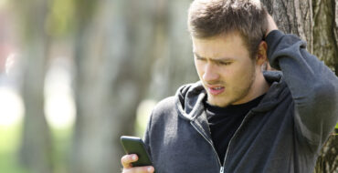 a man dressed in casual clothing, standing in front of a tree, looking confused and concerned as he reads something on his phone