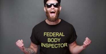a white man with a short beard is wearing a t-shirt that says "Federal Body Inspector". He has a tattoo on his arm and is wearing a black baseball cap and sunglasses. He's got his arms out, fists up, and he's shouting, like he's cheering for something