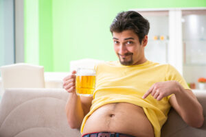A man sitting on a couch has his t-shirt rolled up to expose his belly, which he pointing at with a happy expression