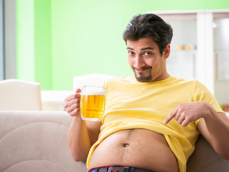 A man sitting on a couch has his t-shirt rolled up to expose his belly, which he pointing at with a happy expression