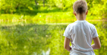 little boy sitting on dock with his back to the camera, looking at a lake surrounded by very green forest