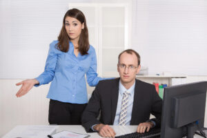 A man is looking at the camera, sitting at a desk in front of a computer, while a woman stands slightly behind him and also looks at the camera while shrugging