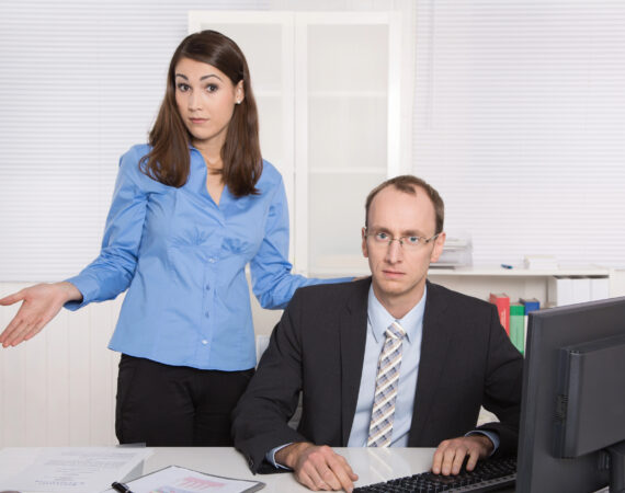 A man is looking at the camera, sitting at a desk in front of a computer, while a woman stands slightly behind him and also looks at the camera while shrugging