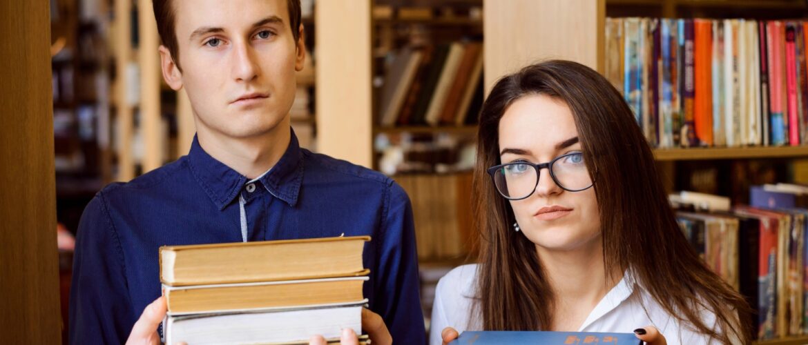 Two teenagers in a library, each holding a stack of three books and looking vaguely unhappy