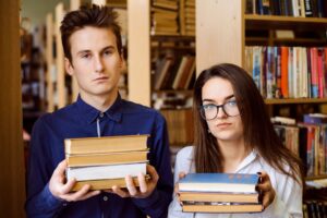 Two teenagers in a library, each holding a stack of three books and looking vaguely unhappy