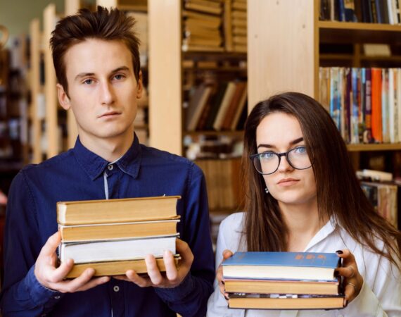 Two teenagers in a library, each holding a stack of three books and looking vaguely unhappy