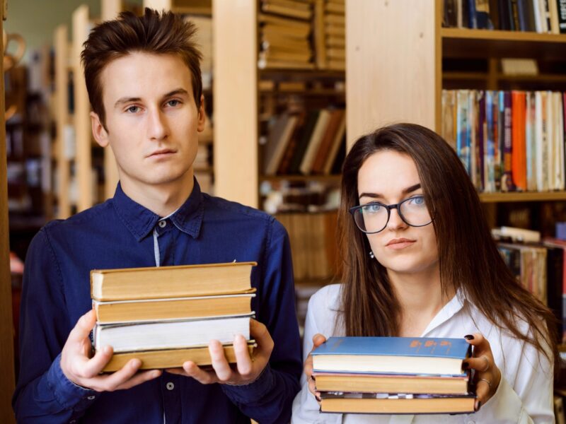 Two teenagers in a library, each holding a stack of three books and looking vaguely unhappy